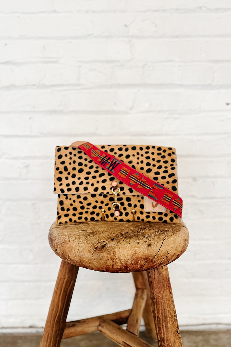 Cheetah leather fold-over clutch with a red personalized webbing crossbody strap attached to it sitting on a stool