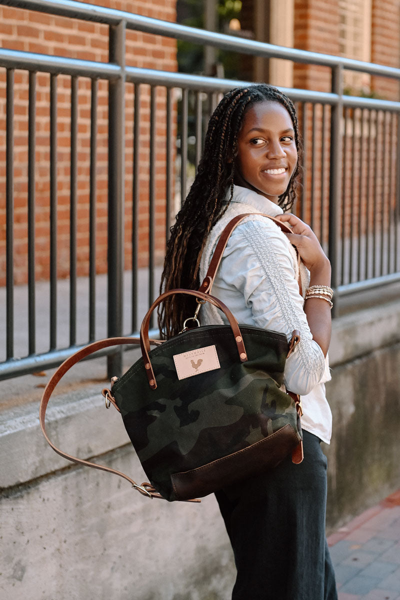 A woman wearing the camouflage backpack with dark brown straps and the meanwhile logo.