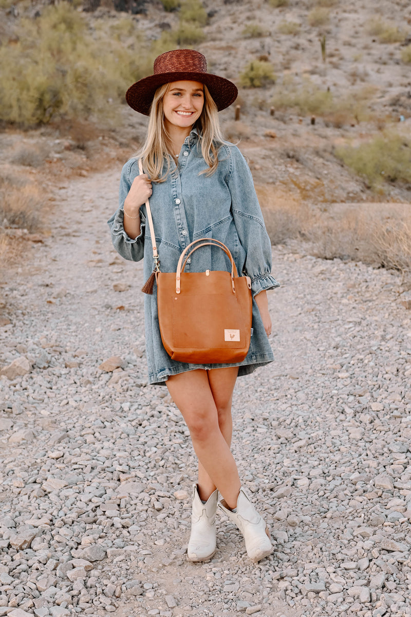 Model reaching into the front pocket of a tan leather purse with cream colored straps.