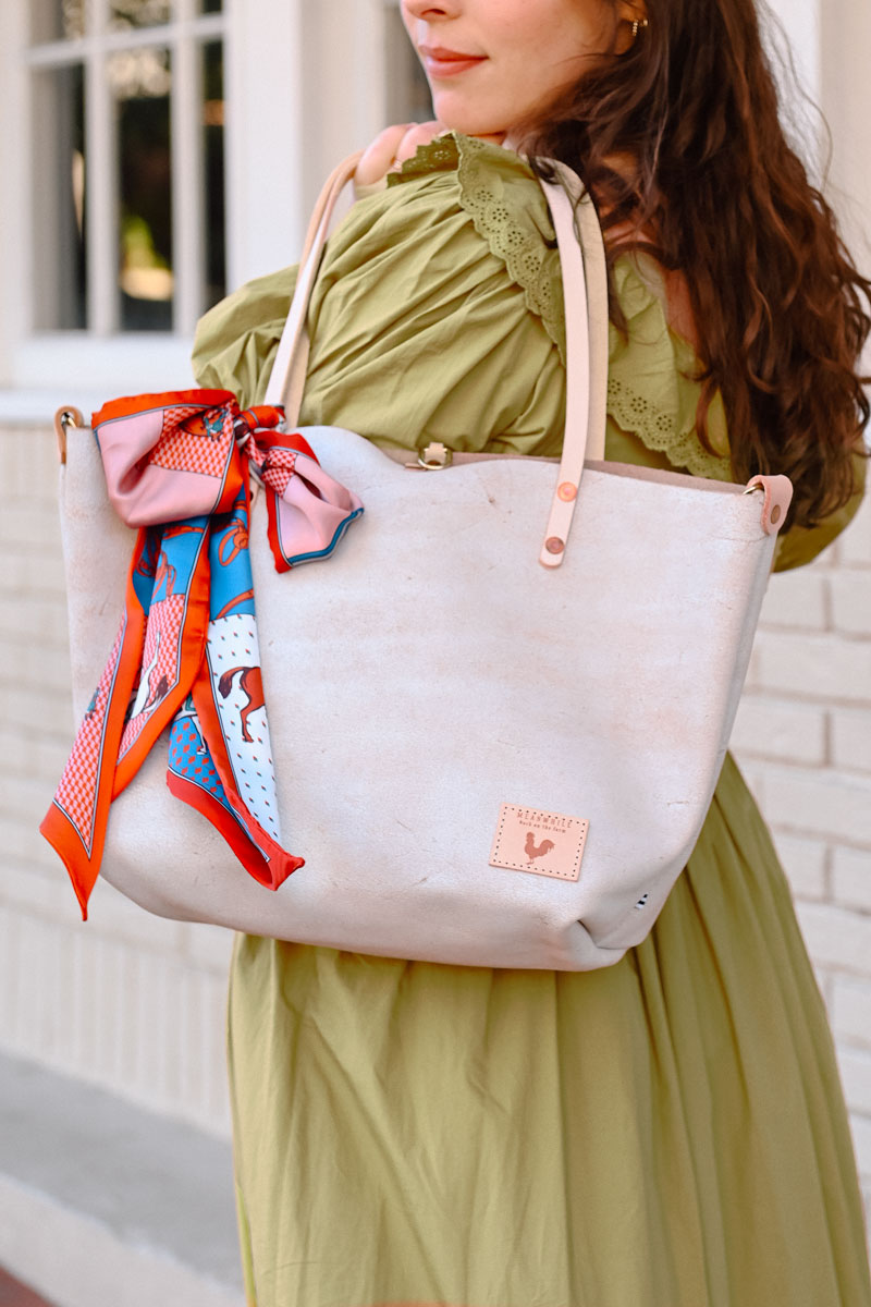 Model in green dress displaying birch white leather tote bag with cream colored handles and accent scarf