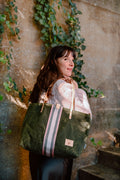 Woman holding large green leather tote thrown over her shoulder. Tote has cream colored straps and accent strip down the center in cream, white and green striping.