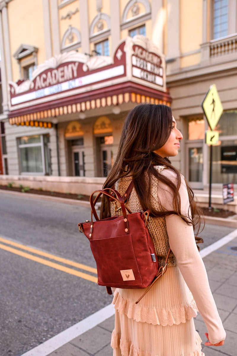 Model crossing the street wearing a ruby red leather bag on her back by it's backpack straps. Bag has a front pocket and handles.