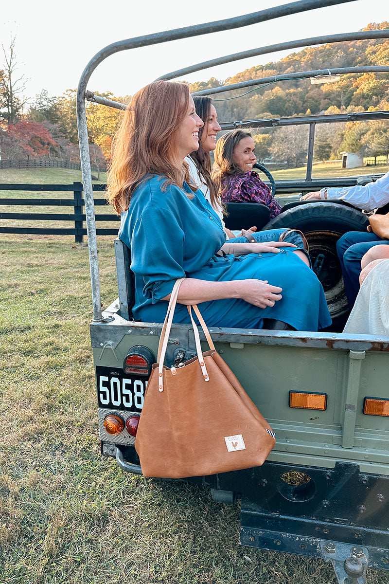 Women in the back of an antique jeep, one is pictured prominently holding a large tan leather tote with cream colored straps.