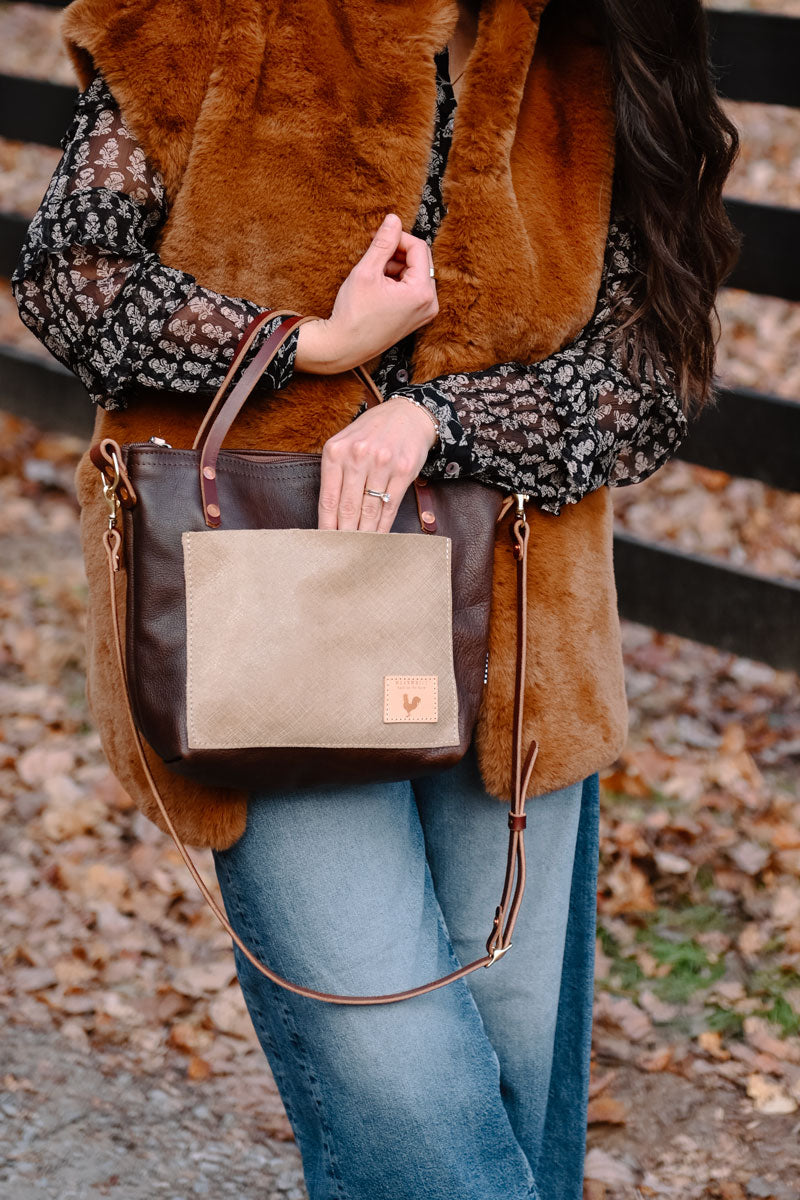 Model reaching into handbag. Handbag is dark leather with a cream colored brushed leather front pocket