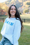 Picture of model in Farm field wearing a white sweatshirt with the words Farm embroidered in blue on the front