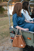 Women in the back of an antique jeep, one is pictured prominently holding a large tan leather tote with cream colored straps.