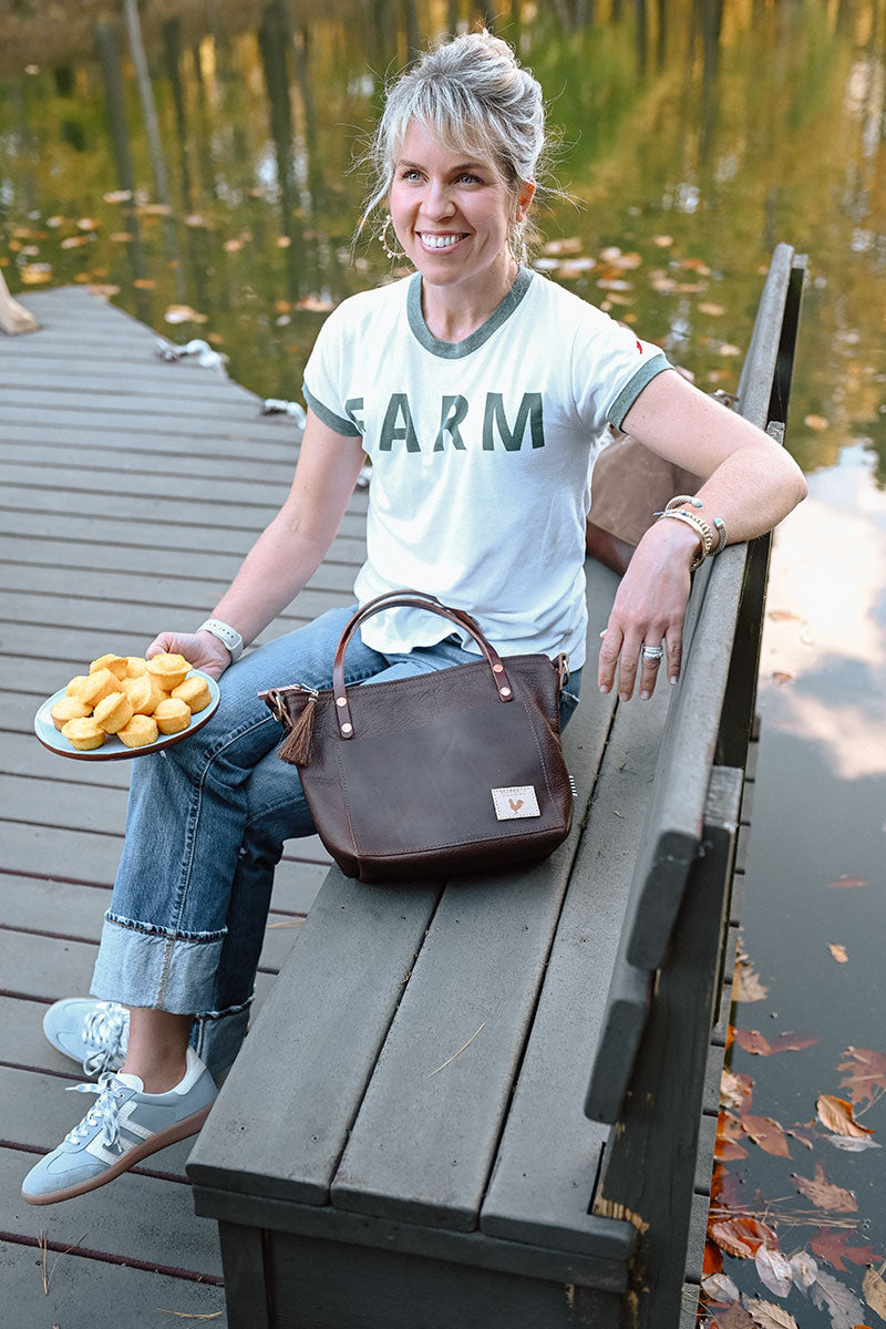 Woman sitting on a dock bench. She is wearing a white Tshirt with green trim and the word Farm embroidered in green on it.  She has a dark brown leather handbag resting on her lap. 