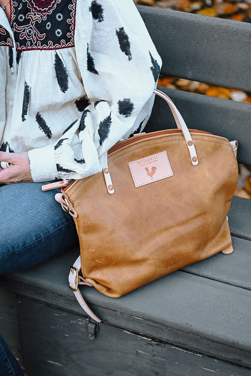 A woman wearing the camel brown backpack with tan straps and the meanwhile logo.