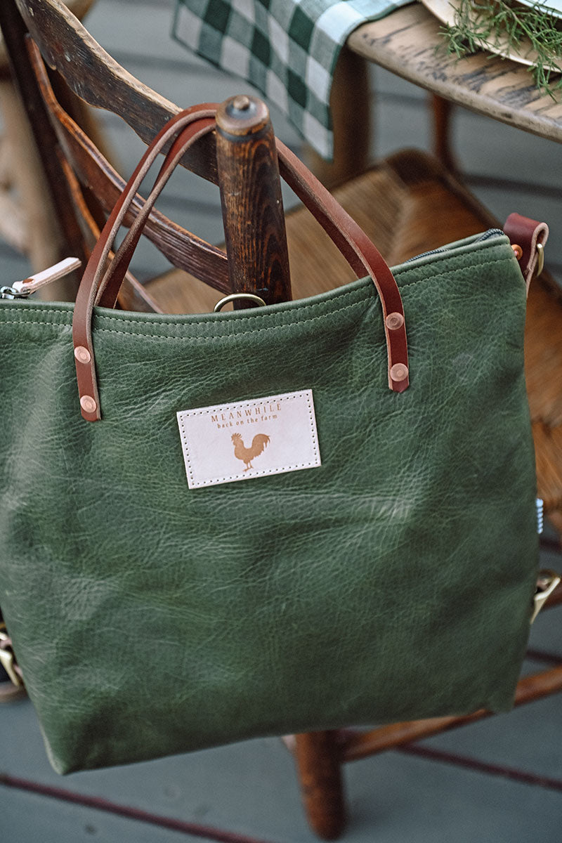 Picture of large dark green leather handbag with brown leather handles hanging on the back of a wooden chair.