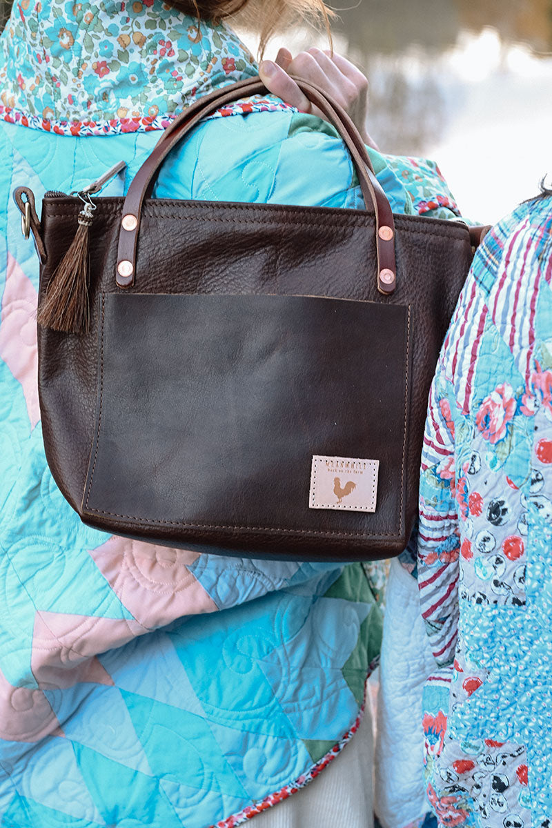 A woman holding a mocha brown short strapped bag with a brown tassel and meanwhile logo.