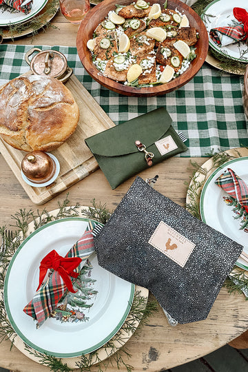 Picture of festively decorated dinner table with seasonal foods and dishes. There are two clutches laying amidst the items on the table. One is hunter green with a clasp and the other is a silver speckled clutch.