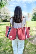 Girl holding two bags behind her: orange leather large tote bag with a red/white stripe in the middle and a matching one with a green/white stripe in the middle 