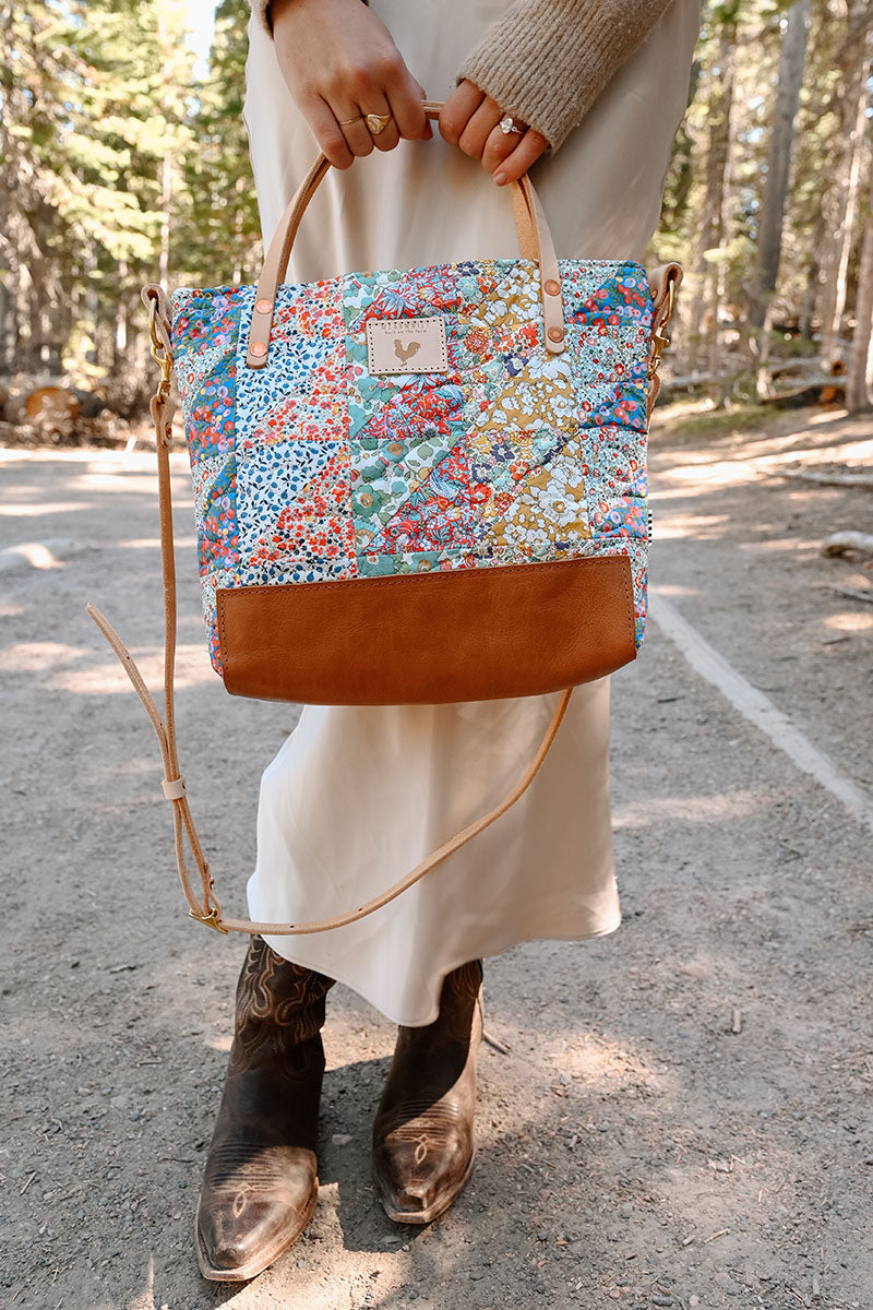 Model holding large handbag in front of her legs. Bag is quilted floral print with cream colored straps and tan leather bottom