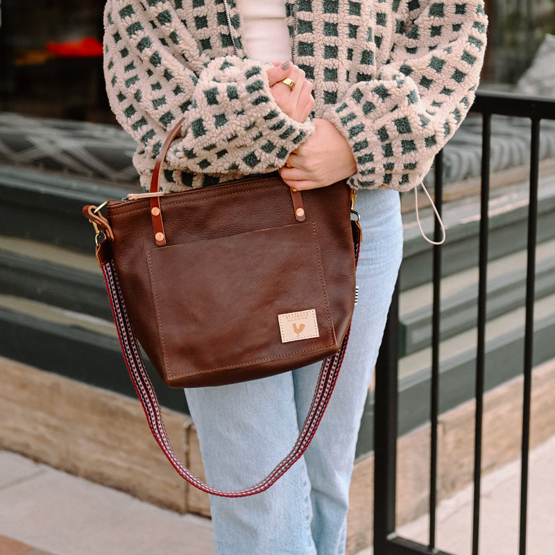 A woman holding a mocha brown short strapped bag with a brown tassel and meanwhile logo.