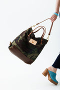 Model walking away holding a large camo print tote with brown leather bottom. Bag has brown straps.