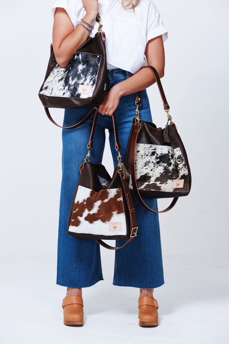 Model holding three different shoulder bags with cowhide accents in varying color options of brown, dark brown and black.