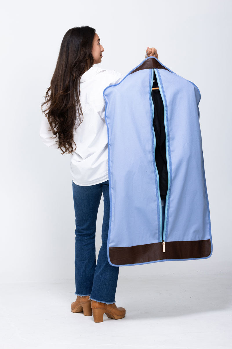 Picture of model holding out a blue garment bag with dark brown leather trim.