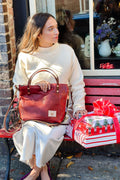 Model waiting on bench and holding a reddish leather bag in her lap. Bag has brown leather handles straps and tan leather shoulder strap. Christmas wrapped packages are sitting next her.