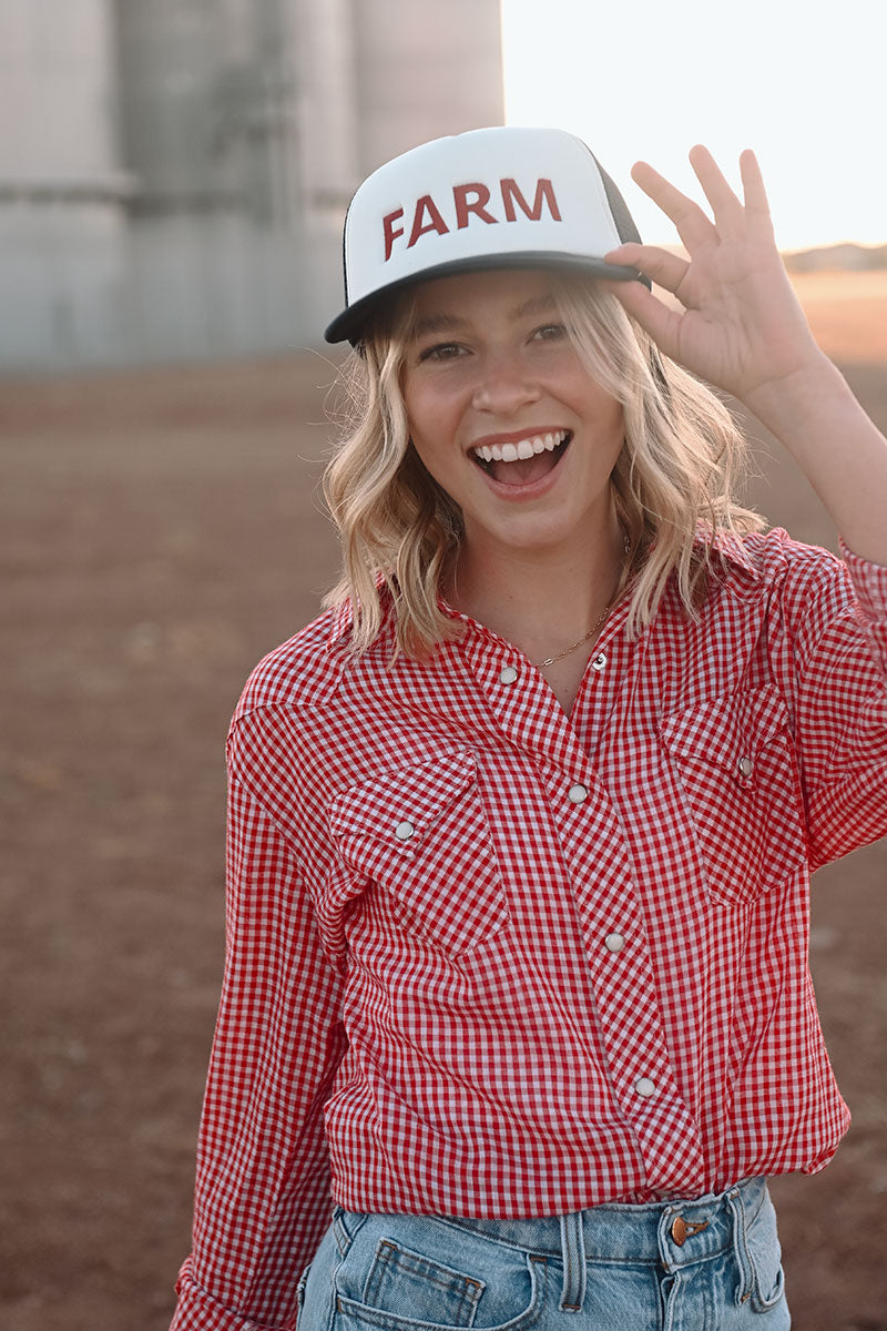 Model in front of silos with a red plaid shirt and jean pants. She is holding the brim of a dark colored trucker hat with the word "Farm" embroidered on the front.