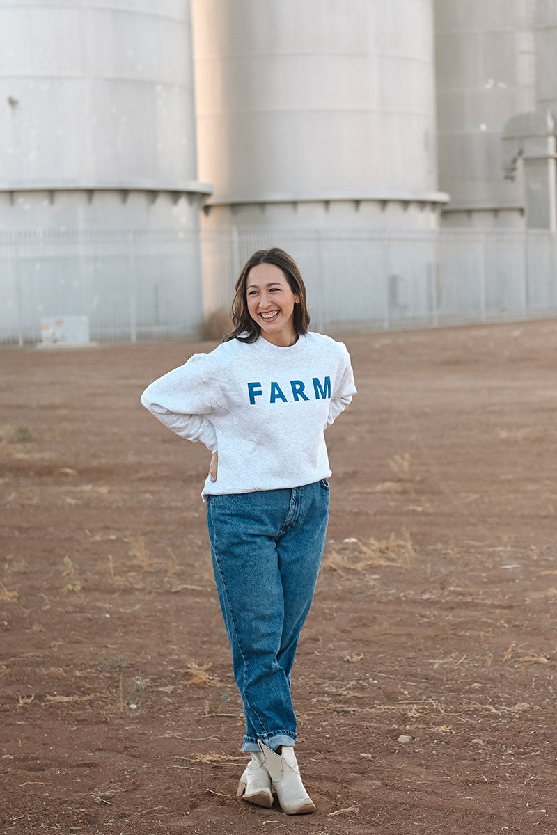 Model in front of silos wearing a white sweatshirt that reads "Farm" and wearing blue jeans.