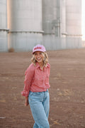 Model in front of silos wearing a red plaid button up and blue jeans. She is wearing a pink hat with the word 