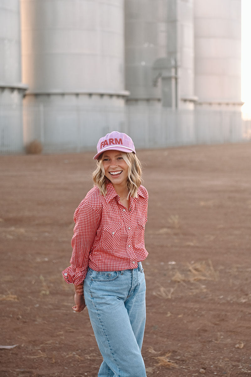 Model in front of silos wearing a red plaid button up and blue jeans. She is wearing a pink hat with the word "Farm" embroidered on it.