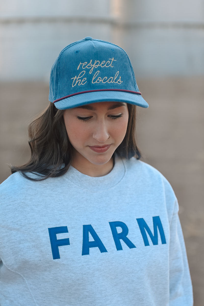 Model wearing a a gray sweatshirt with the word "Farm" printed on the front and wearing a teal colored hat embroidered in cursive script, reading "respect the locals".