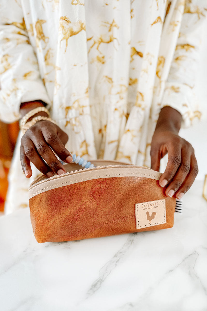 Woman smiling taking a blue/white beaded necklace out of a small light brown leather cosmetic bag