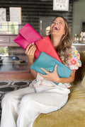 Model laughing joyously holding a collection of large fold over style leather bags in bright pink, teal and salmon.