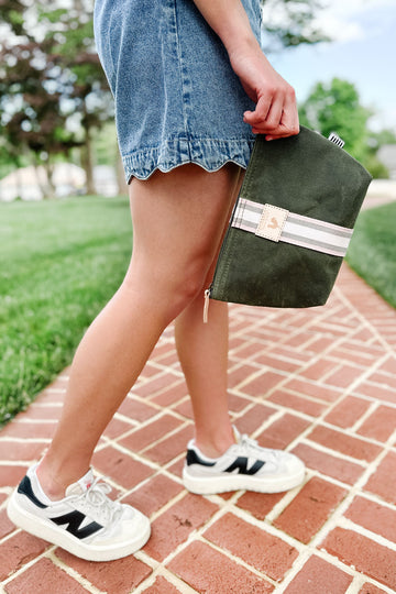 Girl walking on a brick sidewalk holding a small olive green leather makeup bag with a white/gray stripe