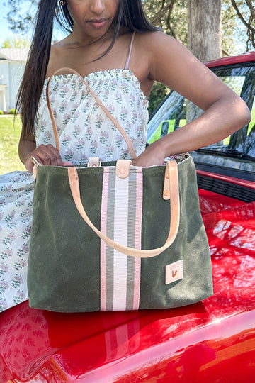 Woman outside sitting on the hood of a red car reaching into an olive green waxed canvas tote with a white/gray/cream stripe in the middle with cream colored straps 