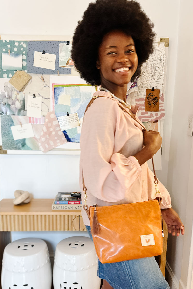 Woman in front of a bulletin board smiling holding a small light brown sling bag in her hands