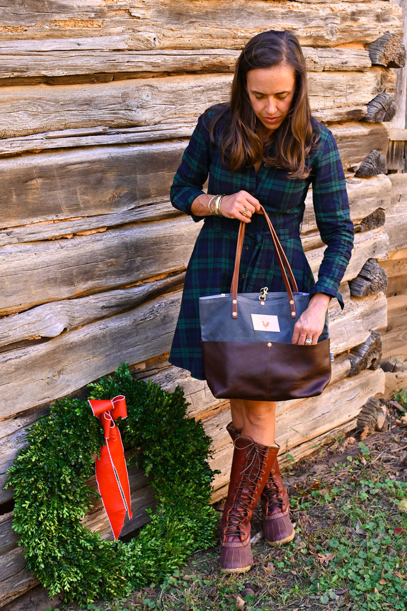 Model looking down into pocket of slate blue utility tote that has a dark brown leather bottom and pockets with brown leather straps.