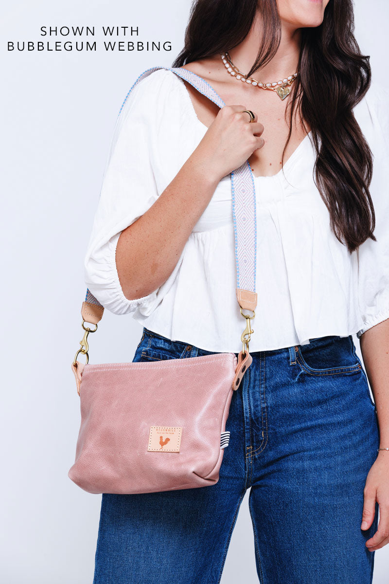 Woman wearing a small pink leather purse with a light blue/yellow/pink webbing crossbody strap attached to it