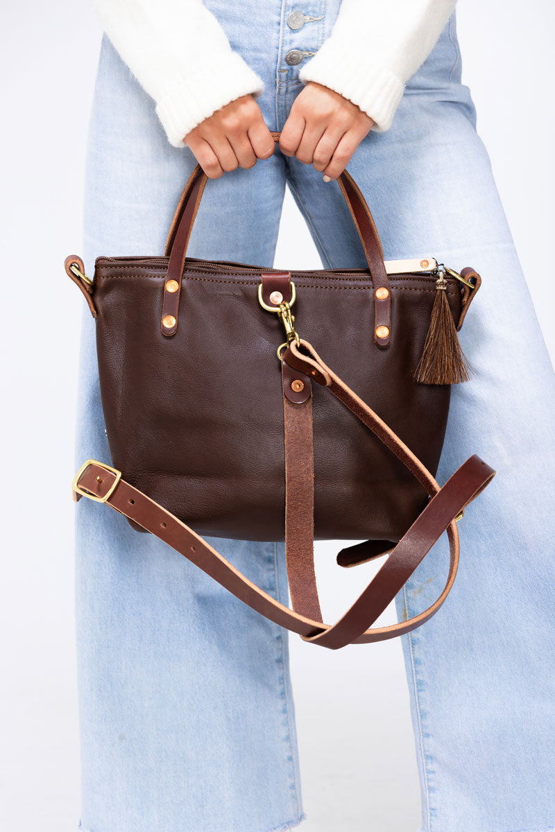 Model wearing a dark brown leather bag by back pack straps.  Bag also has a front pocket and tassel. 