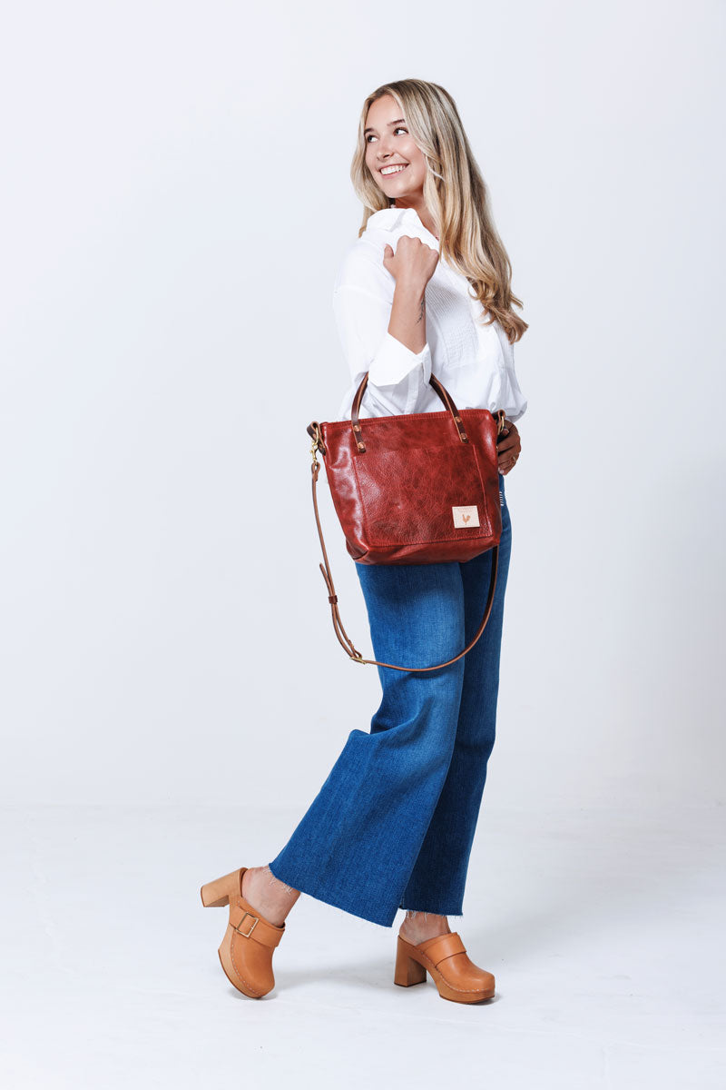 Model looking back over her shoulder, holding a medium sized ruby leather handbag by it's handles with shoulder strap hanging down