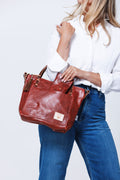 Model placing a ruby leather wallet into the front pocket of a medium sized ruby leather hand bag that she is holding by it's handles