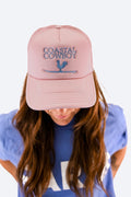 Woman wearing a pink trucker hat that says coastal cowboy in light blue letters with the logo of a rooster on a surfboard