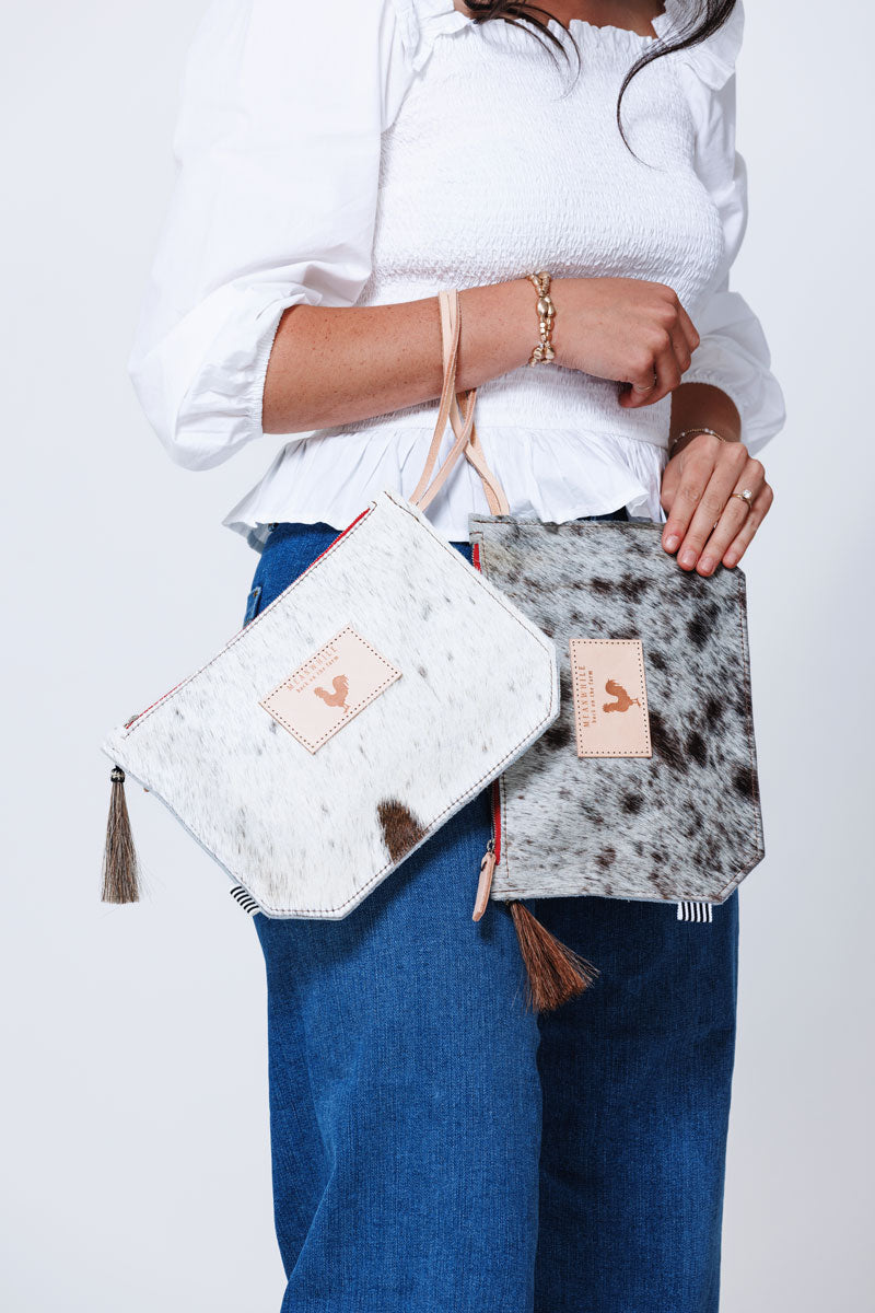 Model displaying two medium sized clutches in cowhide and pink accent tassel and wrist strap and brown accent tassel and wrist strap