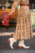 Woman in orange dress wearing white cowgirl boots holding a red/brown leather fold-over clutch and crossbody behind her