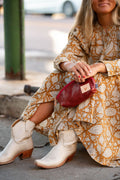 Woman sitting down in an orange dress wearing white cowgirl boots holding a red/brown leather cosmetic bag