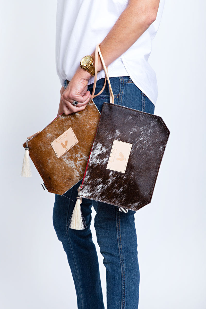 Model displaying two medium sized clutches in cowhide and pink accent tassel and wrist strap and brown accent tassel and wrist strap